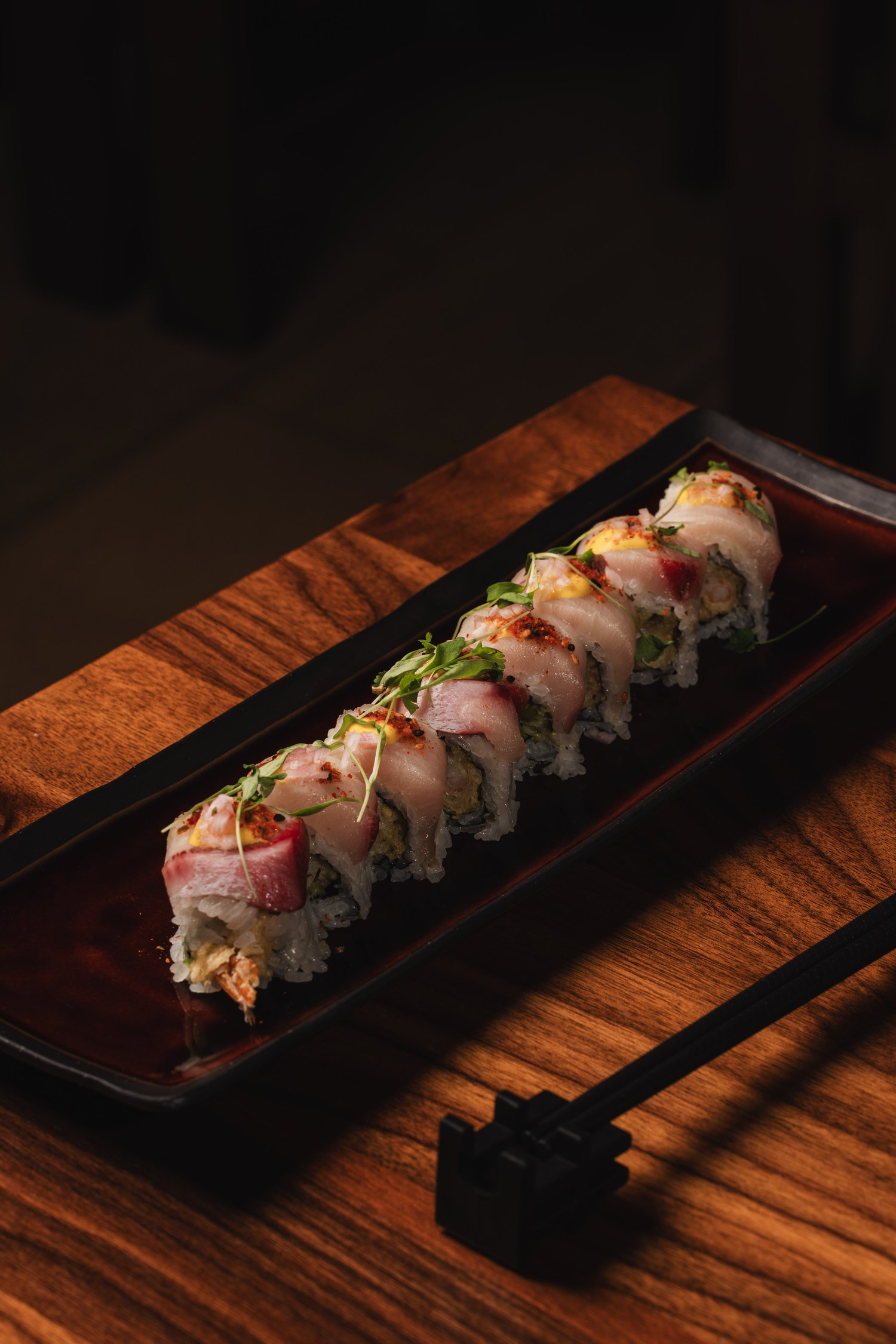 A sushi roll topped with sliced hamachi, chili and micro herbs on a black lacquer tray