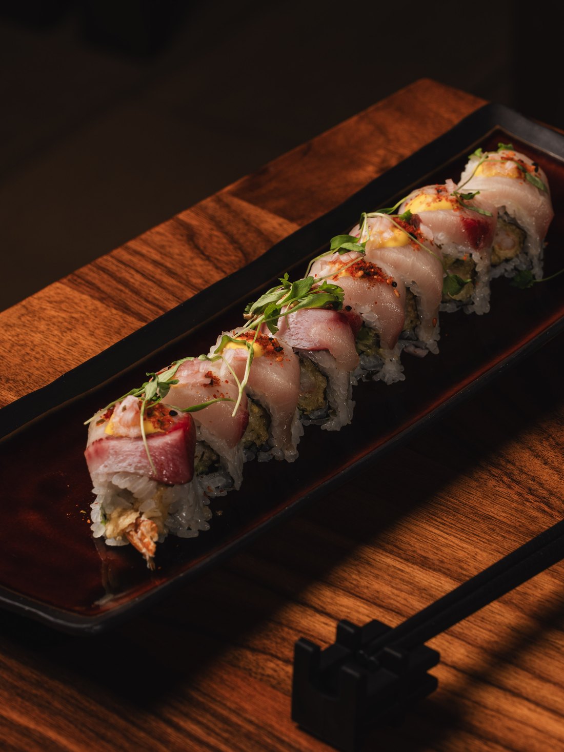 A sushi roll topped with sliced hamachi, chili and micro herbs on a black lacquer tray
